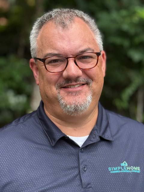 Head-and-shoulders portrait of a smiling man with short gray hair, glasses, and a salt-and-pepper goatee, wearing a dark polo with the SimpleHome logo; photographed outdoors against a softly blurred leafy background.