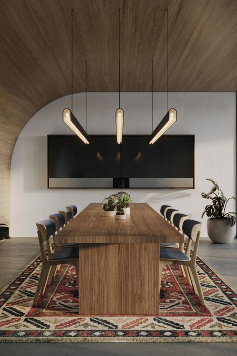 Modern conference room with an arched wood ceiling, three sculptural pendant lights over a long walnut table, eight chairs on a patterned rug, a dual-screen video display on the wall, and a potted plant in the corner.