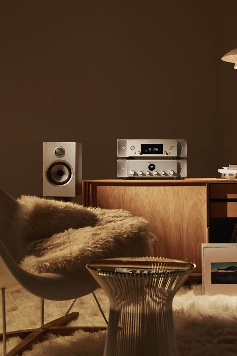 Cozy mid-century listening nook with a sheepskin-draped lounge chair, wire side table, and a walnut console holding a stacked silver stereo receiver and amp; a single bookshelf speaker sits to the left, all warmly lit against a mocha wall and plush rug.