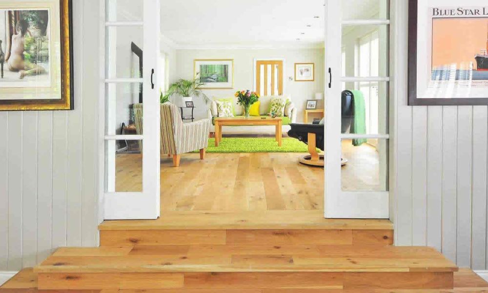 A view into a bright living room with a large window, a green rug, and comfortable seating, seen through open French doors from a hallway with art on the walls.