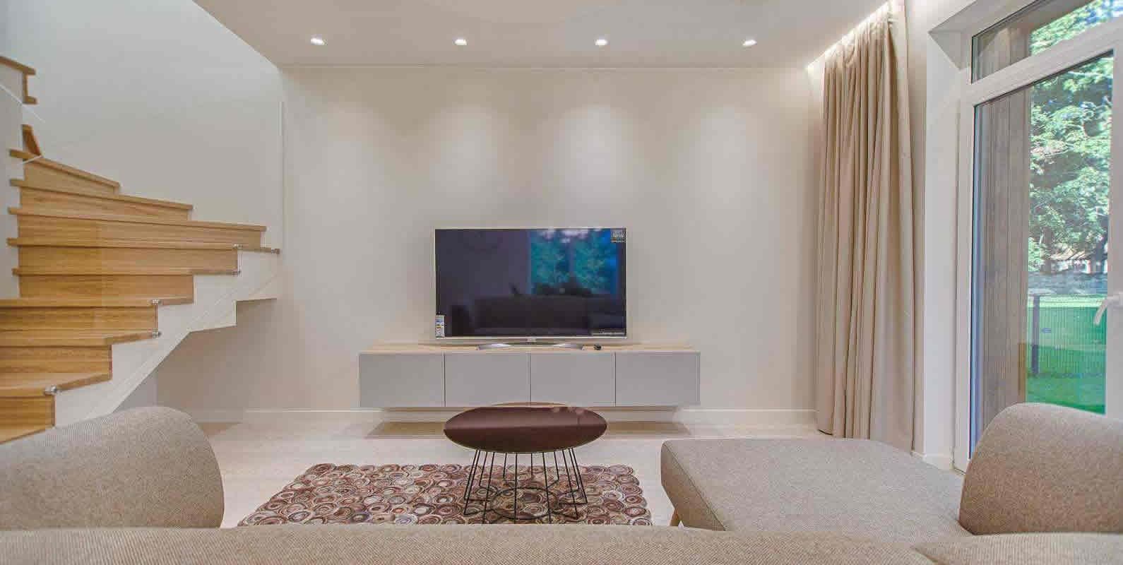 A modern living area with a beige sectional sofa, a flat-screen TV, a wooden staircase, and large windows, offering a bright and open design.