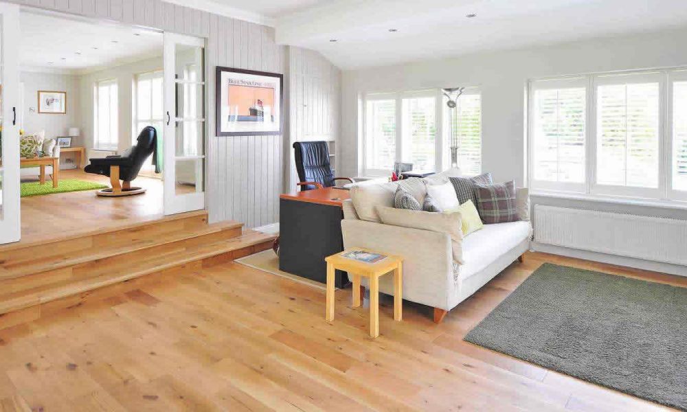 A spacious living area with light wooden floors, a comfortable sofa, a black desk, and large windows with white blinds, offering a bright and inviting atmosphere.