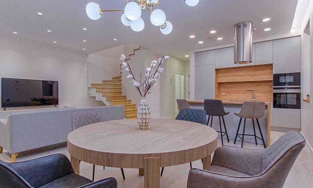 An open-concept living and dining area with a round dining table, gray chairs, a staircase, and a modern kitchen, showcasing a blend of functionality and style.